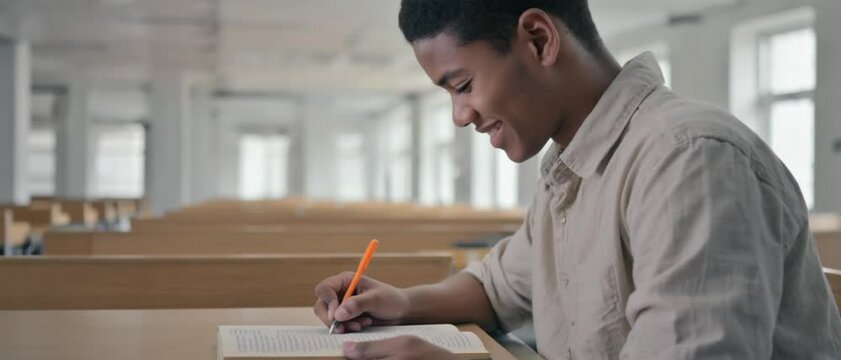 A focused black student smiles while taking handwritten notes alone in quiet classroom rows. Concept of academic dedication and self‑driven learning