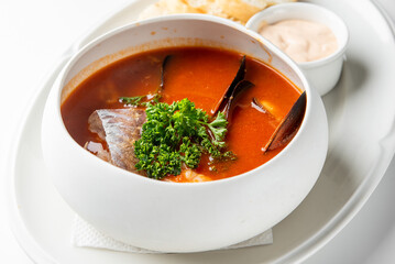 A bowl of tomato-based seafood soup garnished with fresh parsley, served with bread and a small dish of sauce on a white plate.