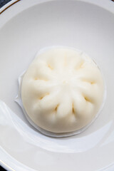 A single, perfectly steamed Baozi or steamed bun is served in a minimalist white bowl. Its soft, doughy texture and signature pleats are highlighted, emphasizing the simple elegance of this popular As