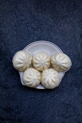 A top-down view of five steamed buns (Baozi) artfully arranged on a white plate with a gold rim, set against a dark, textured gray background. The soft, pleated surface of the buns is prominently feat