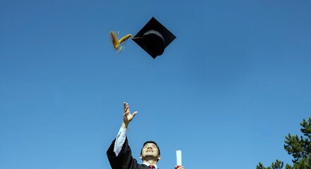 Graduate throwing mortarboard cap against clear blue sky. Student in academic gown celebrating educational achievement with diploma. Graduation ceremony and academic success. University commencement