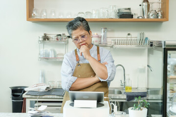 Senior Asian woman cafe owner wearing apron standing with thoughtful expression looking at tablet reflecting on coffee shop management enjoying independent post-retirement family business.