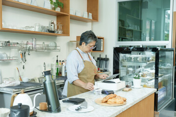 Senior Asian woman cafe owner in apron standing at counter using tablet managing receipts preparing...