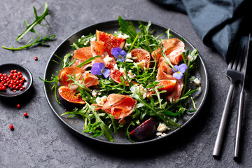 Arugula salad with fresh figs and prosciutto, jamon and edible flowers in a plate. Grey background. Close up.