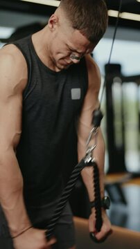 A determined male athlete performs tricep rope pulldowns in a modern gym. He focuses on proper form and muscle engagement during his strength training workout. Building muscle, getting stronger