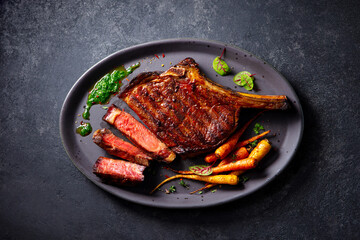 Beef tomahawk steak on a bone with roasted colorful carrots and chimichurri sauce on a plate. Grey background. Close up. Top view.
