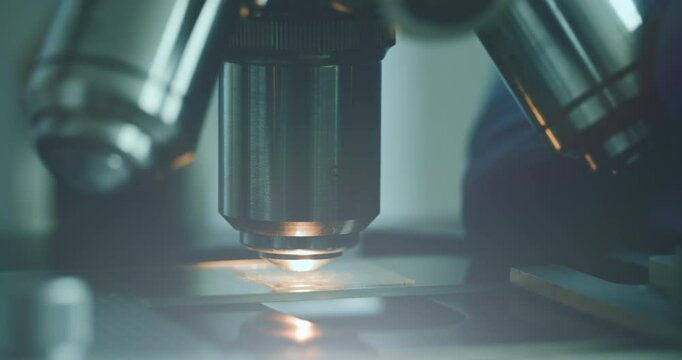 Close-up of biologist gloved hands using microscope in sterile laboratory. Vibrant blue gloves highlight precise scientific work against monochromatic lab backdrop