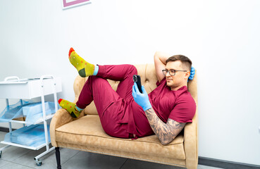 Chill worker in fun socks. A man in scrubs lounges on a couch while checking his phone, wearing...