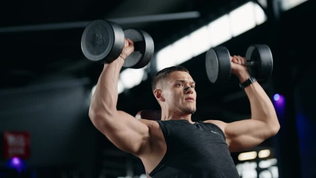 A determined male athlete performs dumbbell shoulder presses in a modern gym. He focuses on proper form and strength training. This video embodies fitness, health, and dedication to exercise.