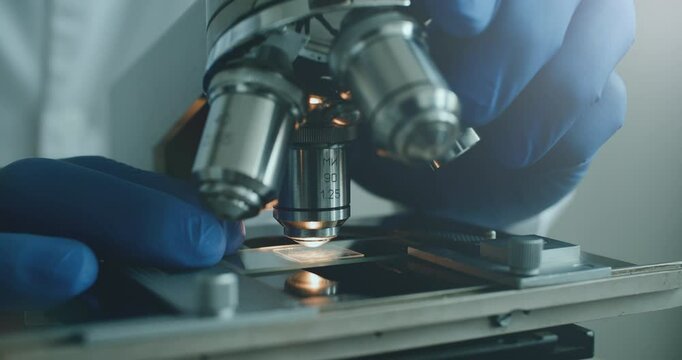 Close-up of biologist gloved hands using microscope in sterile laboratory. Vibrant blue gloves highlight precise scientific work against monochromatic lab backdrop