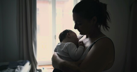 Mother gently cradling baby close while standing near window, sharing intimate and peaceful moment of connection and love in softly lit home environment