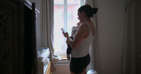 Mother holding her baby while using a smartphone, multitasking in a home environment with soft...