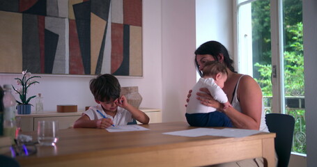 Tired boy rubbing his eye while doing homeschooling work at the table, mother holding baby nearby in a peaceful home setting, highlighting family dynamics and educational focus