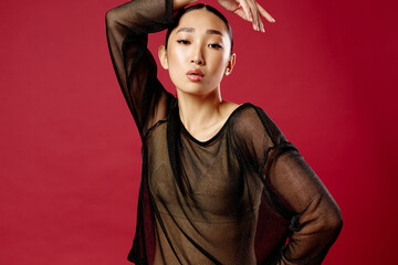 Fashion asian woman in sheer top posing on red background with hands on head and head tilted to side