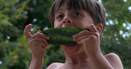 Young boy lifting a slice of watermelon to his face, enjoying a refreshing snack outdoors, highlighting a carefree and joyful moment on a sunny day