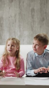 Little blonde girl pupil answers nodding African-American tutor while brother classmate sits on sofa at home English lesson with native speaker slow motion