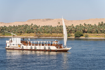 Dahabiya Nile cruise ship sailing alongside a sandy desert with palm trees on the River Nile near Aswan