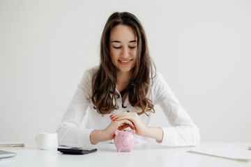 Young woman saving money in a pink piggy bank while sitting at a desk with a calculator and cup during daylight hours