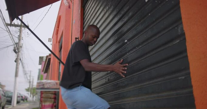African American man pulling down metal shutter to close small grocery store at dusk with shopping carts and crates visible inside