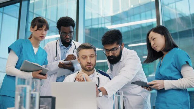 Diverse group of doctors gathered around head physician at laptop, Indian man pointing at screen, others holding tablets and clipboards. Medical team collaborating during intensive case review.
