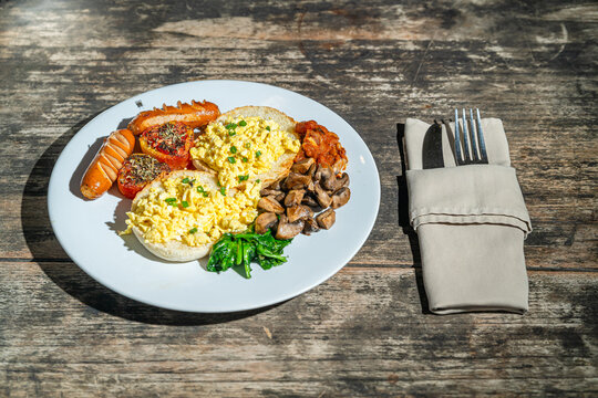 Big breakfast with bread toast, scrambled egg, sausages, roasted tomato, baked bean, sauted mushroom, and wilted spinach on white plate. Isolated by wooden table. Food Photography