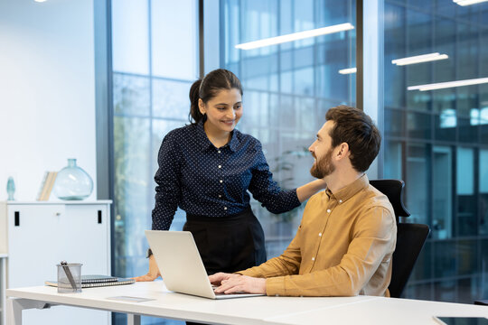 A woman smiles while placing her hand on a man's shoulder as he works on a laptop in a bright office setting.