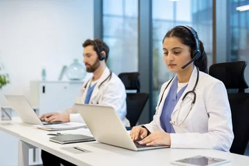 Fotobehang Two doctors are seen in an office setting, wearing headsets and using laptops. They may be providing remote medical consultations. © Liubomir