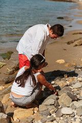 A mother and her teenage son closely inspect the texture of a large stone on a sunny beach filled with rocks and surrounded by calm water.