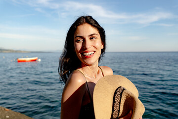 Tourist enjoying a sunny summer vacation at the beach, holding her straw hat while gazing out at...