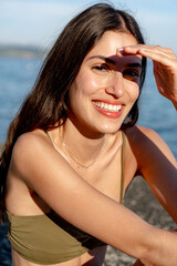 Young woman with long brown hair shielding her eyes with her hand from the sun, smiling and...
