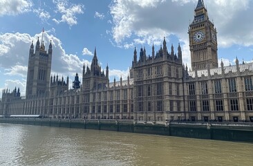 Fototapeta premium Historic British parliament building alongside a river, under a partly cloudy sky. Tall towers and intricate architectural details are visible