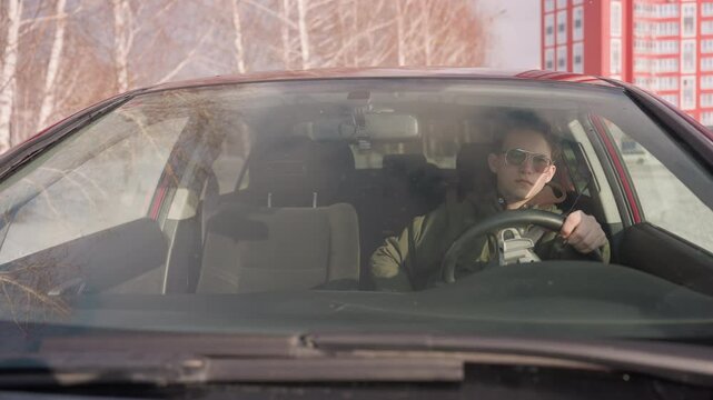 teenager wearing winter jacket seen through windshield sitting in parked car holding steering wheel with left hand while right hand rests, seatbelt unfastened, red building and snowy landscape