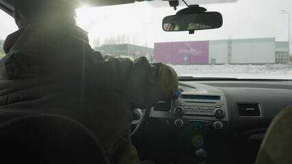 back view of man cleaning steering wheel with blue cloth inside parked car while truck and small car drive past on snowy road beyond window under soft winter light early morning calm