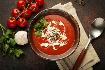 Fresh tomato soup is served in a bowl with cream, seeds and herbs, accompanied by cherry tomatoes, garlic and parsley on a brown background with a spoon and napkin