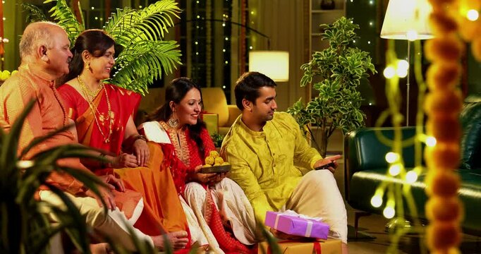 Clicking Diwali selfie with laddu, Indian family with young girl, senior mother in saree, old father and husband in traditional wear celebrating Hindu festival at decorated home with diya rangoli