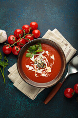 Fresh tomato soup is served in a bowl with cream, seeds and herbs, accompanied by cherry tomatoes, garlic and parsley on a blue background with a spoon and napkin