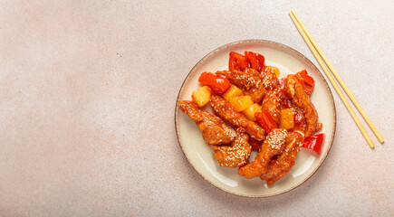 Asian delicious sweet and sour chicken garnished with sesame seeds, pineapple chunks, and bell pepper slices on a plate with chopsticks on light beige background