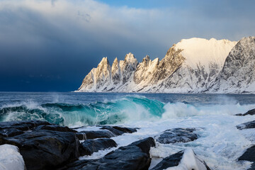 Die Okshornan auf Senja in Norwegen zur goldenen Stunde, im Licht leuchtend vor einem aufziehenden dunklen Sturmhimmel, während hohe Wellen im Vordergrund das aufgewühlte Meer bestimmen