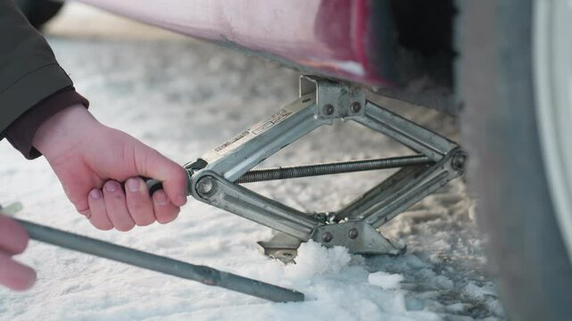 Close up of car mechanic using spanner to turn jack screw and increase height while working under parked vehicle on snowy ground, hands adjusting metal parts