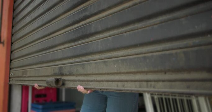 Latina woman closing metal shutter of grocery store from inside at end of workday, lowering storefront gate with effort while finishing daily routine