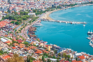 Fototapeta premium Aerial view of Alanya's harbor with colorful pirate ships docked along the promenade and nearby beach. The bustling coastal area offers a beautiful panorama of the city and the Mediterranean Sea