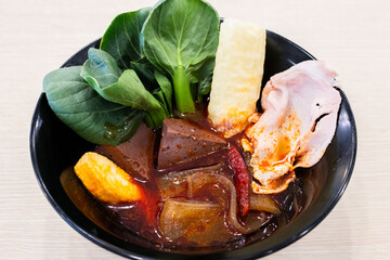A vibrant bowl of spicy Asian soup featuring pork blood cubes, leafy greens, chili peppers, sliced pork, fish cake, and crispy fried tofu skin