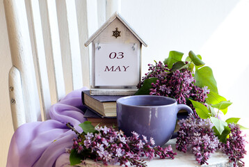 Calendar for May 3: numbers 03, word May in English on decorative calendar, cup of tea, lilac branches on chair