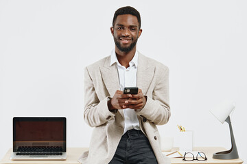 Smiling man using smartphone in bright office space, wearing casual blazer, standing in front of desk with laptop, glasses, and lamp. Office workspace, technology, communication, work from home