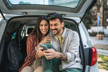 Young adult couple sit on car trunk and use mobile phone at street