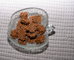 A glass dish shaped like a leaf holds an assortment of crunchy chocolate pieces. The light reflects off the surface, creating a tempting display on a soft, textured background