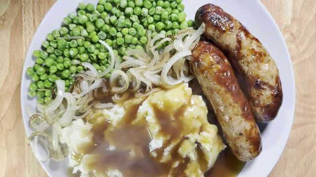 Closeup overhead shot of steaming hot gravy over mashed potato on a dinner plate with cooked sausages, peas and fried onions.