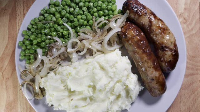 Closeup overhead shot of steam rising from a dinner plate of hot cooked sausages, peas, fried onions and mashed potato.