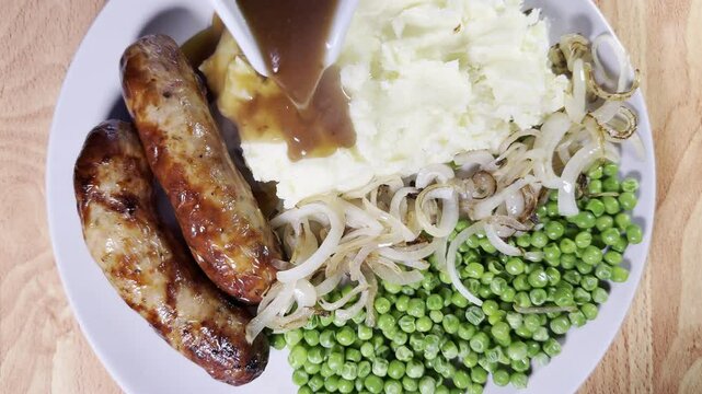 Closeup overhead shot of steaming hot gravy slowly pouring over mashed potato on a dinner plate with cooked sausages, peas and fried onions.