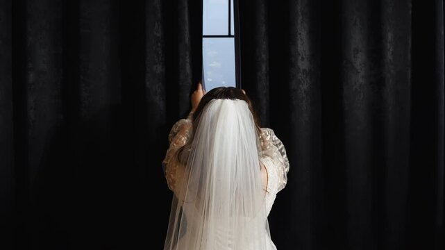 Back view of a bride in a white dress and with veil on the head, opening black curtains to the sides in front of a window with her female hands in room. Symbolic wedding tradition. No face